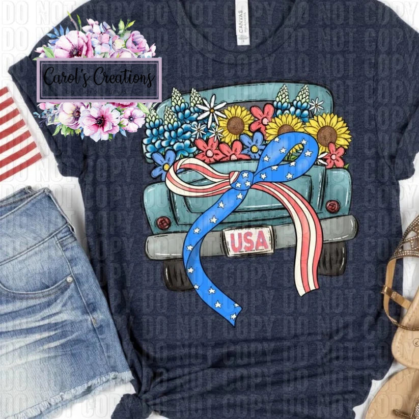 Navy blue t-shirt with a graphic of a truck carrying flowers and a blue ribbon, with 'USA' on the license plate, next to denim shorts and an American flag.