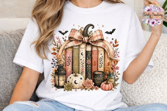 A woman wearing a white graphic t-shirt with a fall-themed book bundle design, sitting on a gray couch. The graphic features stacked books, a large bow, pumpkins, a lantern, and autumn leaves.