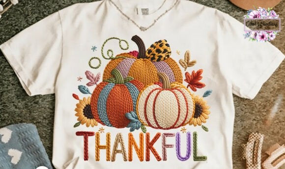A white short-sleeve V-neck T-shirt laid flat on a textured gray surface, featuring a colorful autumn graphic with embroidered-style pumpkins, sunflowers, leaves, and the word 'THANKFUL' in multicolored letters.
