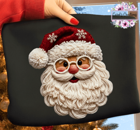 A hand with red nail polish holds a black square fabric item featuring a large embroidered Santa Claus face with a red hat, white beard, and round glasses. A red sleeve is visible at the top left, and a decorated Christmas tree and window are in the background.