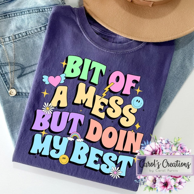 Purple t-shirt with colorful text 'Bit of a mess but doin my best' on a denim background.