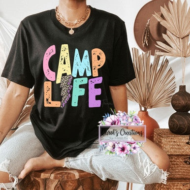 Person wearing a black t-shirt with 'CAMP LIFE' in colorful letters, sitting in a decorative setting.