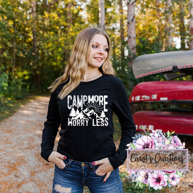 Woman wearing a black sweatshirt with 'Camp More Worry Less' text, standing outdoors with trees and a red vehicle in the background.