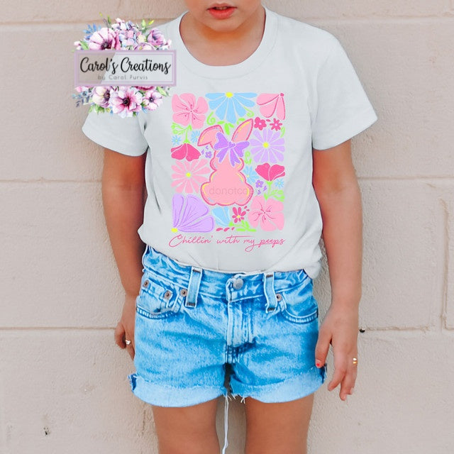 Child wearing a white t-shirt with a colorful bunny design and denim shorts, standing against a light-colored wall.