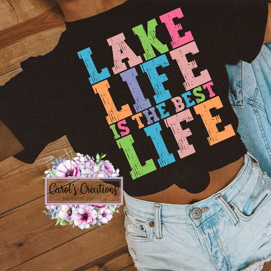 Person wearing a black t-shirt with colorful text 'LAKE LIFE IS THE BEST LIFE' on a wooden floor.