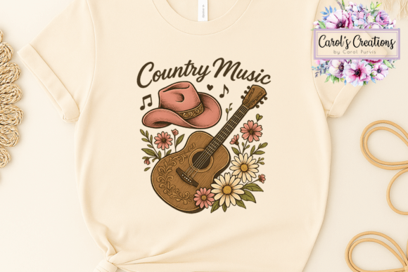 A cream-colored short-sleeve t-shirt featuring a 'Country Music' graphic with a pink cowboy hat, brown acoustic guitar, and pink and white flowers. The shirt is displayed in a flat-lay arrangement on a light beige background with decorative twine and wooden props.