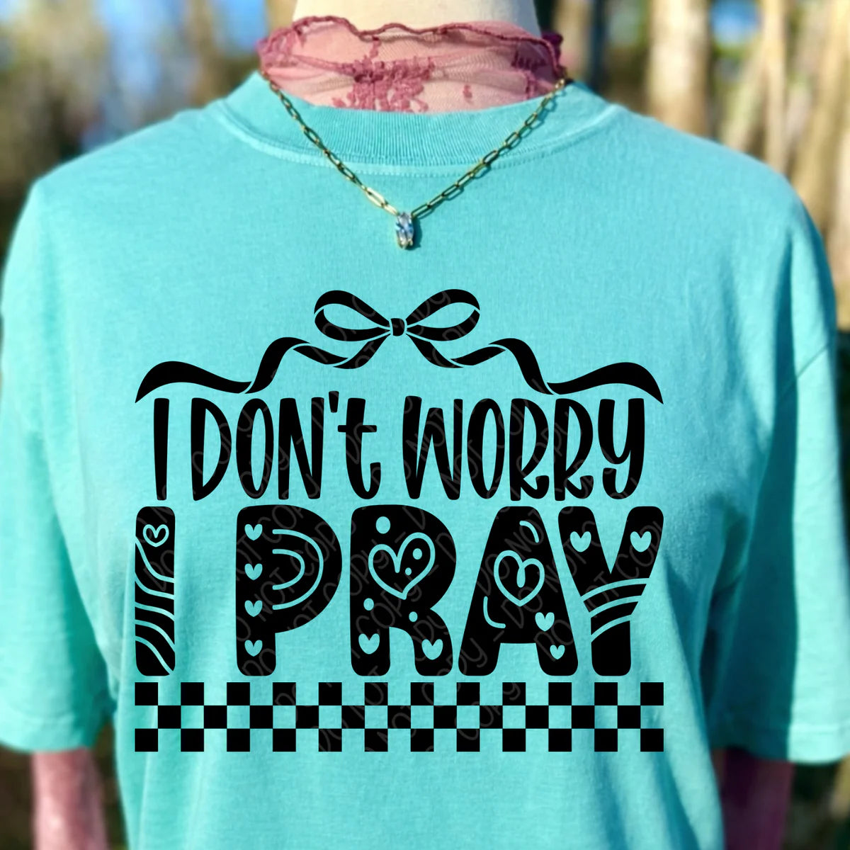 Turquoise t-shirt with black text 'I Don't Worry Pray' on a blurred natural background