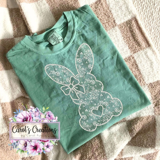 Green t-shirt with white lace bunny design on a textured surface, branded 'Carol's Creations'.