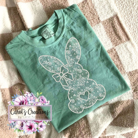 Green t-shirt with white lace bunny design on a textured surface, branded 'Carol's Creations'.