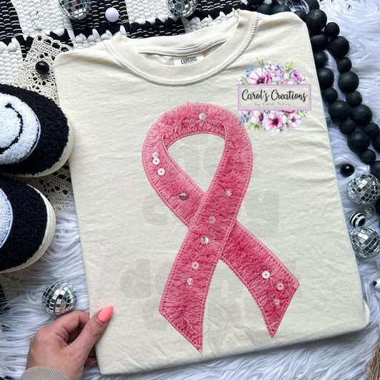 White sweatshirt with pink ribbon design held by a hand, surrounded by decorative items.