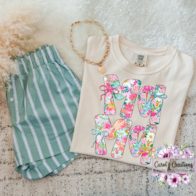 Beige sweatshirt with floral 'MOM' letters, green striped shorts, and gold necklace on a textured surface.