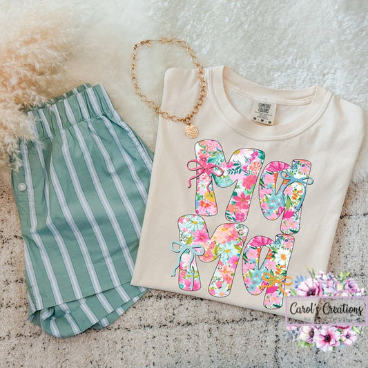 Beige sweatshirt with floral 'MOM' letters, green striped shorts, and gold necklace on a textured surface.