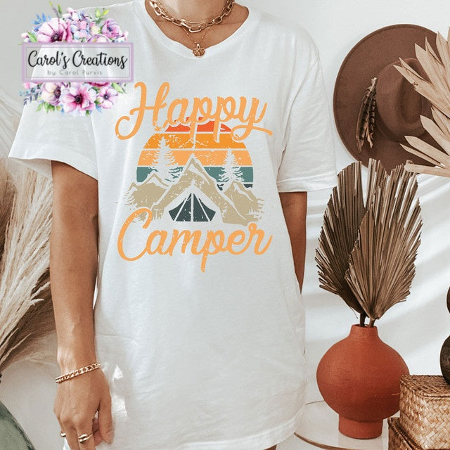 White t-shirt with 'Happy Camper' design, worn by a person, with decorative elements like a hat and vase in the background.