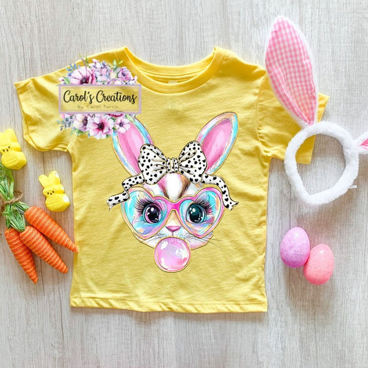 Yellow t-shirt with a colorful bunny design, surrounded by Easter-themed items on a wooden surface.