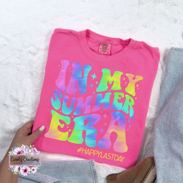 Pink t-shirt with colorful text 'IN MY SUMMER ERA #HAPPYLASTDAY' held by a hand on a white surface.