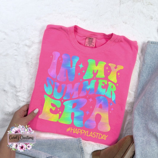 Pink t-shirt with colorful text 'IN MY SUMMER ERA #HAPPYLASTDAY' held by a hand on a white surface.