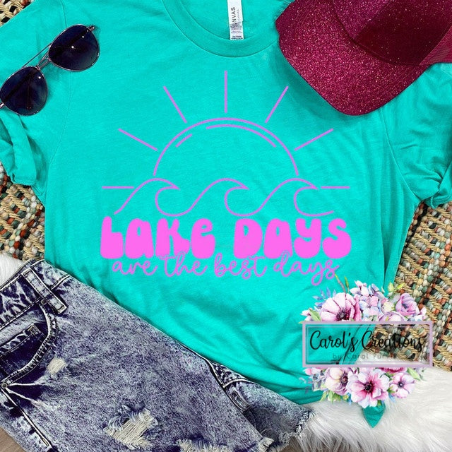Turquoise t-shirt with pink 'Lake Days are the best days' text, sunglasses, hat, and floral decorations on a textured surface.