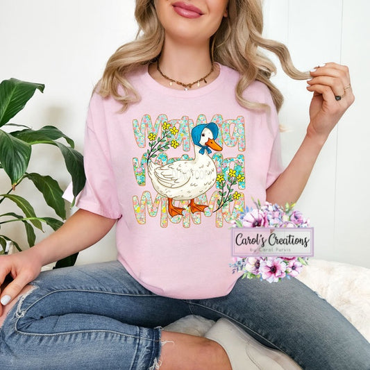 Person wearing a pink t-shirt with a colorful duck graphic and text, sitting on a white surface with a plant in the background.