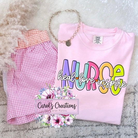 Pink shirt with 'NURSE heal love inspire' text, pink checkered shorts, and necklace on a textured surface.