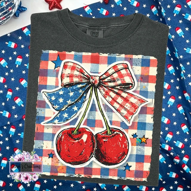 Black t-shirt with a graphic of cherries and a bow on a checkered background, placed on a festive surface.