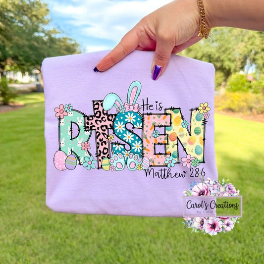 Hand holding a white pillow with 'He is Risen' text and Easter designs outdoors.