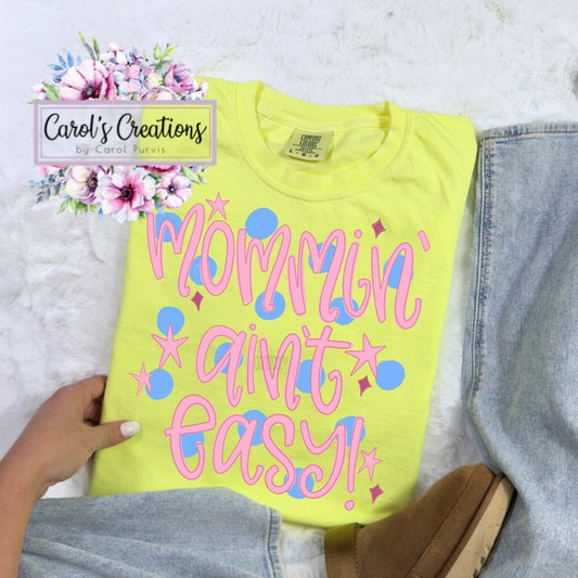 Yellow t-shirt with 'Mommie ain't easy' text held by a person on a light gray background.