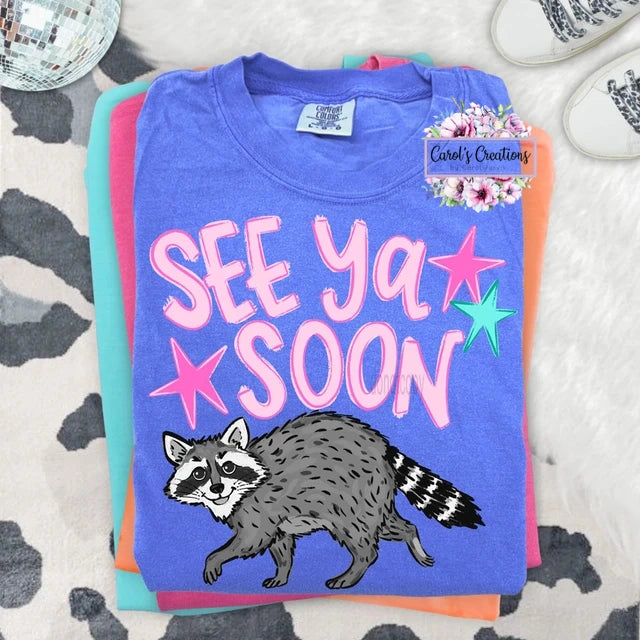 Folded t-shirt with raccoon illustration and 'See Ya Soon' text on a patterned surface