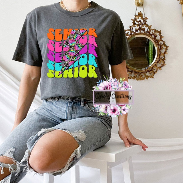 Person wearing a gray t-shirt with colorful 'SENIOR' design, sitting on a white surface.