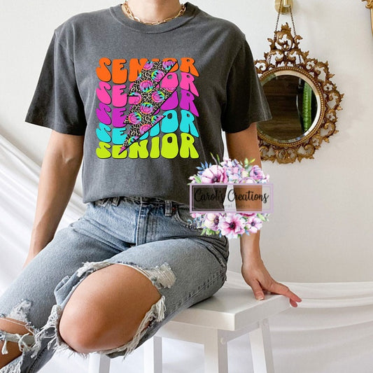 Person wearing a gray t-shirt with colorful 'SENIOR' design, sitting on a white surface.