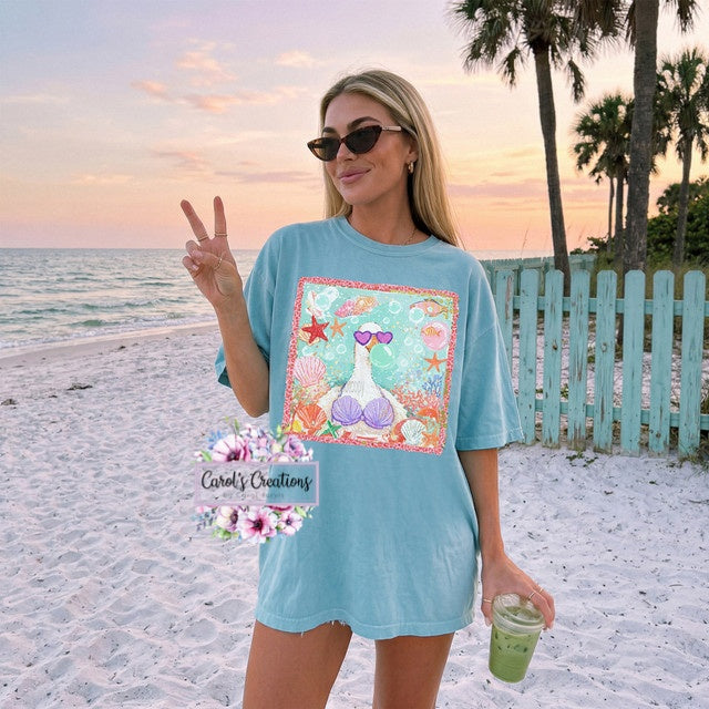 Woman on a beach wearing a blue t-shirt with a mermaid graphic, holding a drink and making a peace sign.
